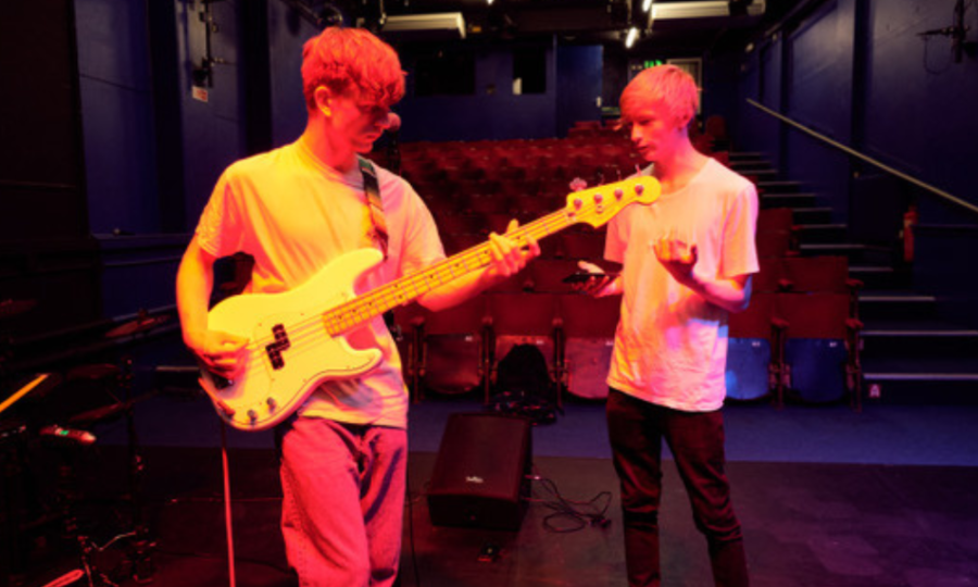 To young men performing on the Seagull Theatre stage. Singing and playing the guitar. The stage is lit up in orange and red tones.