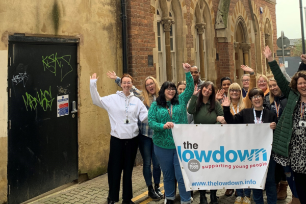 The lowdown team are standing outside their building in Nortampton. They are holding a lowdown banner and celebrating their Youth Investment Fund grant, waving their arms and smiling.