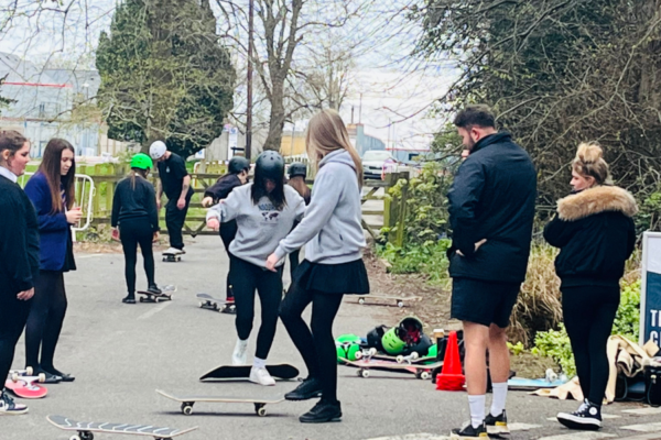 A group of young people outside practising their skateboarding skills together. The are in a park setting, with lots of trees around them.