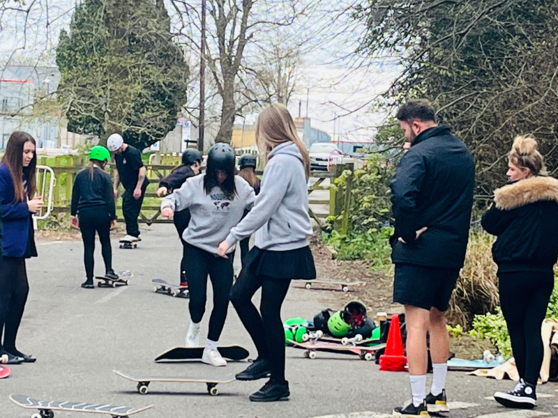 A group of young people outside practising their skateboarding skills together. The are in a park setting, with lots of trees around them.